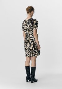 Lyhythihainen musta ja beige kuviollinen mekko, jossa on kapea vyötärö, yhdistettynä polviin ulottuviin mustiin teksturoituihin saappaisiin. Yksinkertainen tausta.