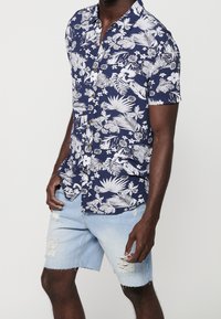 Homme portant une chemise bleu marine à manches courtes à boutons avec un imprimé floral blanc et un short en jean bleu clair usé.