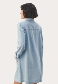 Svetlo modra denim obleka s dolgimi rokavi, gumbnimi manšetami in stranskimi žepi. Značilna je s kragnico in šivom čez hrbet.