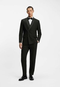 Abito da smoking nero doppiopetto con rever in satin, abbinato a una camicia bianca e a un papillon nero. Presenta una vestibilità su misura e scarpe lucide.