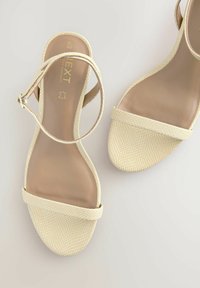 Beige öppna sandaler med en strukturerad meshöverdel, tunn ankelrem och en slät beige innersula, med en minimalistisk design.