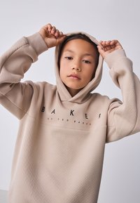 Smėlio spalvos džemperis su kapišonu, su ribinėmis detalėmis, juodu "BAKER" logotipu ir laisvu siluetu. Audinys atrodo minkštas ir tekstūruotas.