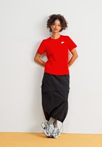 Tricou roșu cu mânecă scurtă și un logo alb mic, asortat cu o fustă neagră lungime până la glezne, ce are buzunare, și adidași gri și albaștri.