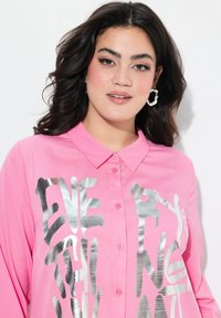 Rosa Kragenhemd mit einem glänzenden silbernen Schriftzug-Design, mit Knöpfen auf der Vorderseite und langen Ärmeln. Glatte Stofftextur.