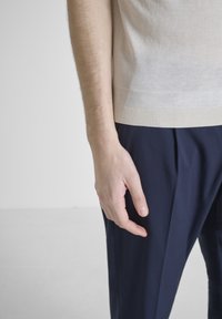 Maglia a maglia di un beige chiaro con orlo a costine, abbinata a pantaloni blu scuro con pinces. Mano visibile che riposa sulla coscia, texture della pelle liscia.