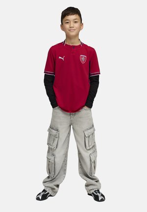 Poika seisoo käsien taskuissa, hänellä on yllään punainen urheilupaita, mustat pitkähihat alla, harmaat cargo-housut ja mustavalkoiset tennarit.