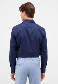 Donkerblauwe overhemd met lange mouwen, gemaakt van een gladde stof en met een puntkraag, gecombineerd met lichtblauwe op maat gemaakte broeken met knoopsluiting op de achterzakken.