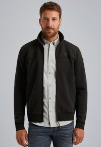 Chaqueta negra con cremallera y una sección superior texturizada, puños elásticos y un ligero cuello alzado, sobre una camisa de botones clara.