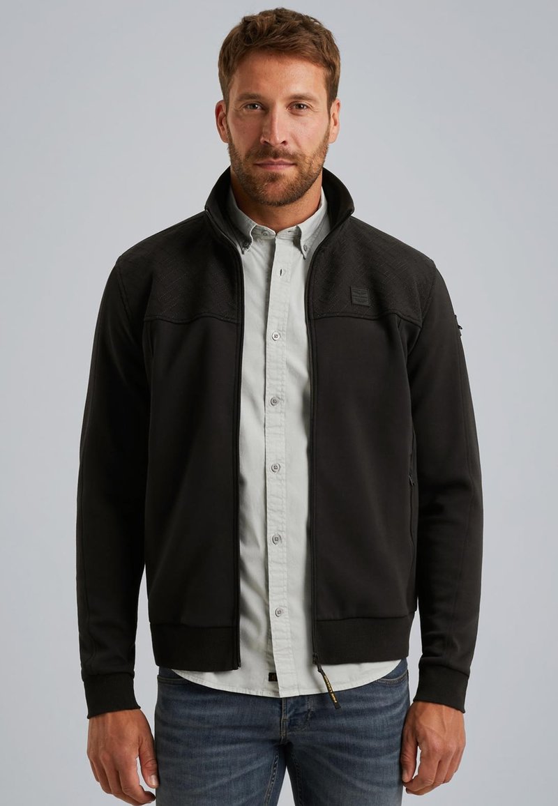 Chaqueta negra con cremallera y una sección superior texturizada, puños elásticos y un ligero cuello alzado, sobre una camisa de botones clara.