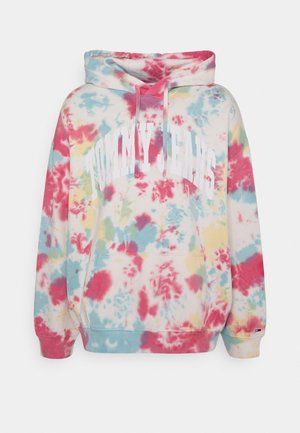 Person trägt pastellfarbenen Batik-Hoodie und passende Sweathose mit weißen Sneakers, steht drinnen in der Nähe einer Treppe und Wand.