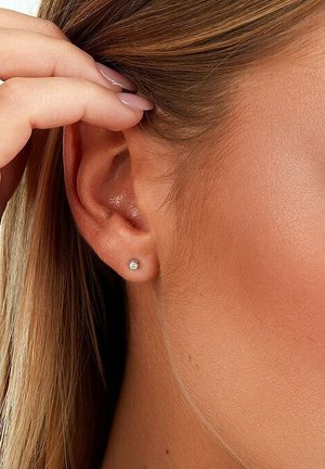 Silberner Steckerohrring mit einem einzelnen runden klaren Edelstein, in einem minimalistischen Design gesetzt. Hintergrund in Hauttönen mit herausgehobenem Ohr.
