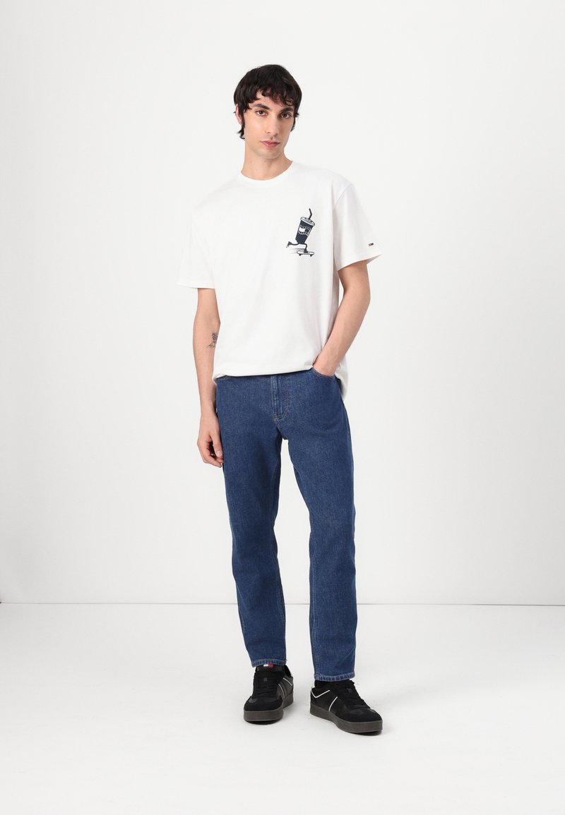 Camiseta blanca con un estampado gráfico negro en el pecho, combinada con jeans rectos azules. El modelo lleva zapatillas negras. Fondo blanco liso.