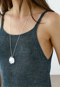 Débardeur en maille bleu sarcelle foncé avec des bretelles fines, présentant une texture lâche et respirante. Un collier pendentif argenté repose autour du cou.