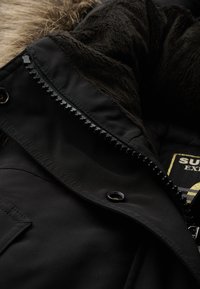 Czarna kurtka z pluszowym, brązowym futerkiem wewnątrz; ozdobiona solidnym zamkiem błyskawicznym oraz metalowymi akcentami w okolicy kołnierza i kieszeni.