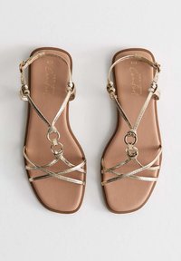 Gouden metalen sandaaltjes met een platte bruine zool en een ronde teen, voorzien van decoratieve ronde hardware en crisscross banden.