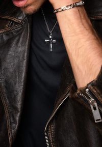 Brun läderjacka med dragkedjor, svart t-shirt, silverkorshalsband och en kedjearmband. Närbild av överkropp och accessoarer.