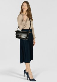 Borsa a mano in pelle nera con chiusura a patta, dettagli in metallo dorato e tracolla regolabile, abbinata a una camicetta beige e una gonna plissettata blu navy.