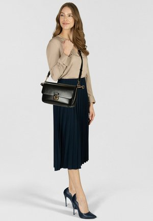 Borsa a mano in pelle nera con chiusura a patta, dettagli in metallo dorato e tracolla regolabile, abbinata a una camicetta beige e una gonna plissettata blu navy.