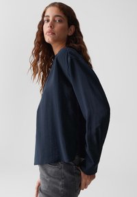 Giovane donna con lunghi capelli castani ondulati, che indossa una blusa larga di un blu navy scuro e jeans grigi, in piedi contro uno sfondo chiaro e semplice.
