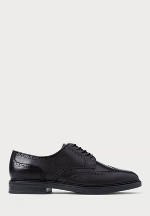 Must mustad brogue-kingad mustast nahast, kaunistatud perforatsioonidega, lipsuga kinnitusega ja madala virnastatud kontsaga heledal hallil taustal.