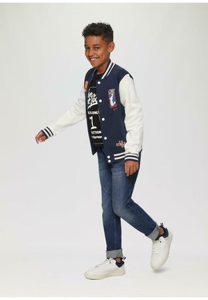 Marineblauw en witte varsityjack met geborduurde patches, gecombineerd met een zwart graphic T-shirt, blauwe jeans en witte sneakers met marineblauwe accenten.