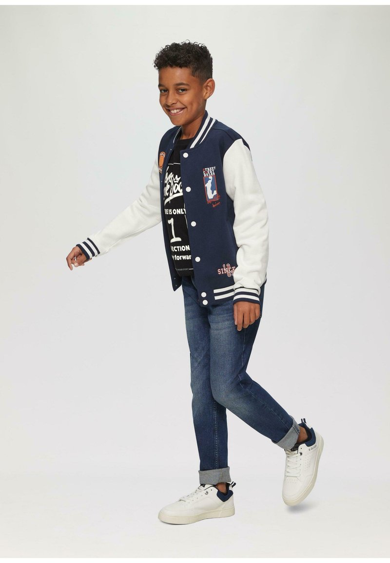Marineblauw en witte varsityjack met geborduurde patches, gecombineerd met een zwart graphic T-shirt, blauwe jeans en witte sneakers met marineblauwe accenten.