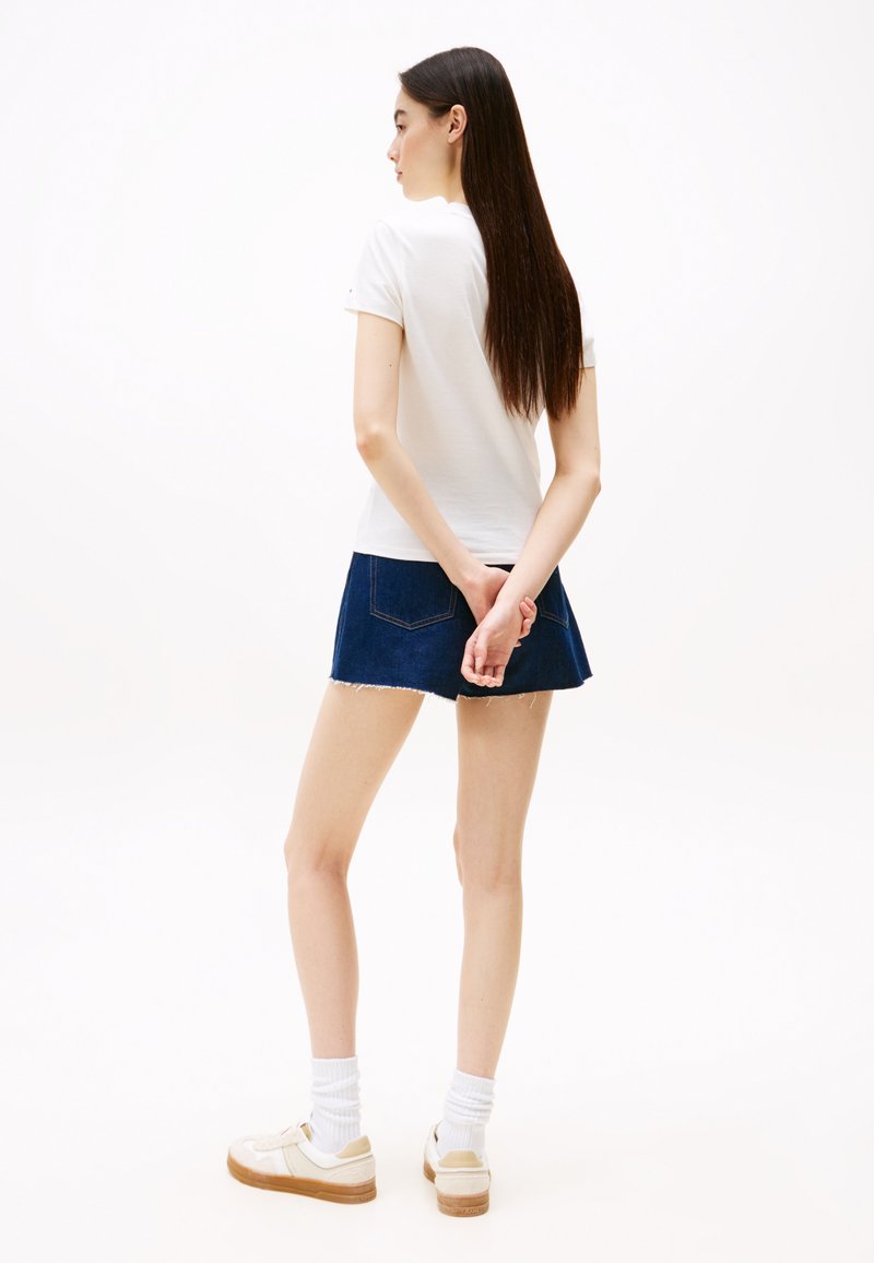 Camiseta blanca de manga corta combinada con una falda de mezclilla azul, que presenta un dobladillo deshilachado. La modelo lleva zapatillas blancas con acentos beige y calcetines blancos.