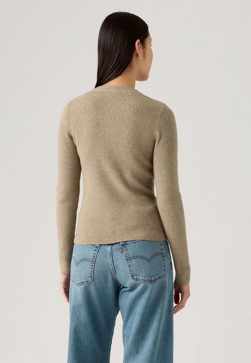 Donna con i capelli lunghi e scuri che indossa un maglione beige a coste a maniche lunghe e jeans in denim blu a vita alta, in piedi contro uno sfondo semplice.