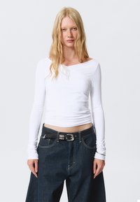 Top bianco a maniche lunghe con scollatura asimmetrica, abbinato a jeans larghi di denim scuro con una cintura nera e fibbia argentata.