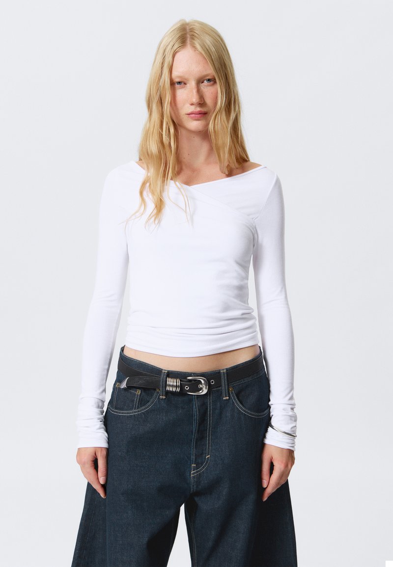 Top bianco a maniche lunghe con scollatura asimmetrica, abbinato a jeans larghi di denim scuro con una cintura nera e fibbia argentata.