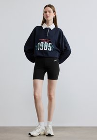 Μπλε σκούρο φούτερ με λευκό γιακά και λογότυπο, συνδυασμένο με μαύρο εφαρμοστό σορτς. Το μοντέλο φοράει λευκά αθλητικά παπούτσια με γκρι λεπτομέρειες και λευκές κάλτσες.