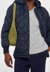 Marineblå quiltet pufferjakke med hætte, grå kabelstrikket sweater under, og en grøn ruskindskrosstaske. Slank pasform og lynlåslommer.