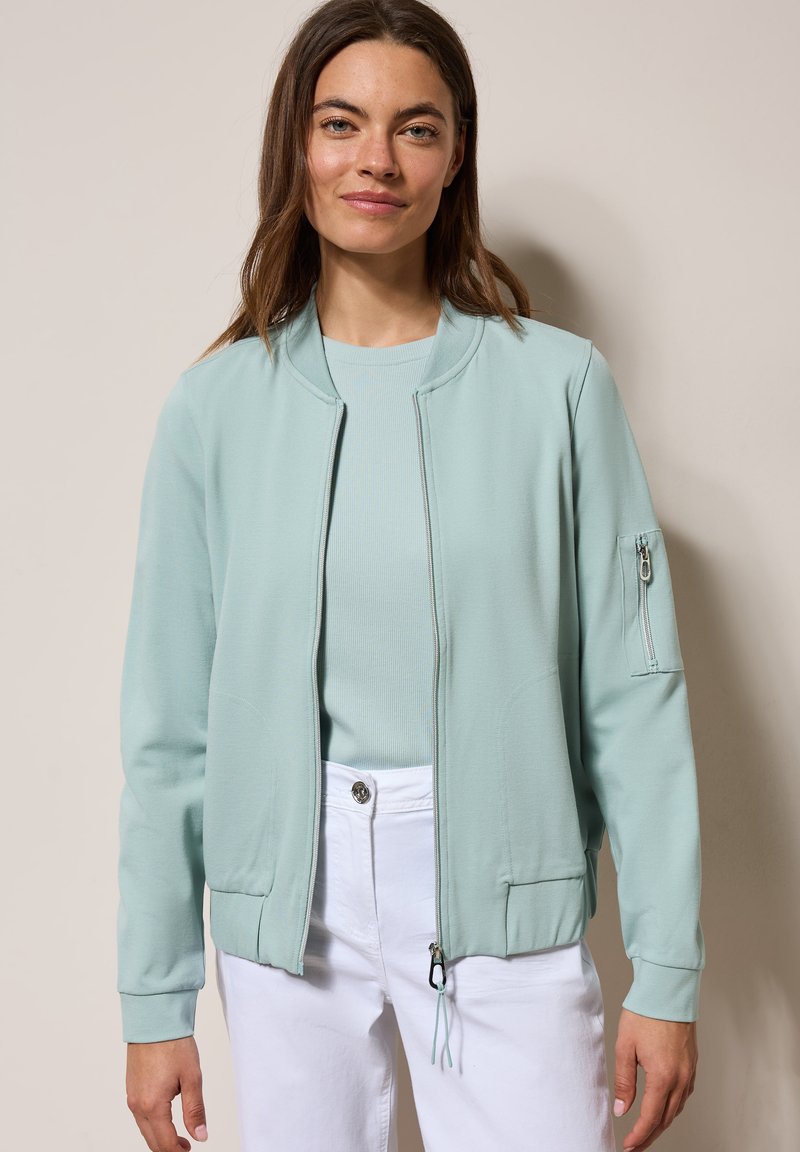 Helle grüne Bomberjacke mit Reißverschluss, gerippten Bündchen und einer Reißverschlusstasche am Ärmel, getragen über einem passenden hellblauen Oberteil.