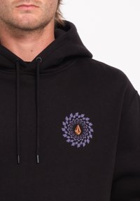 Zwarte hoodie met een cirkelvormige grafische print in paars en oranje, met trekkoorden en een zachte textuur. Opvallend draaipatroon aan de voorkant.