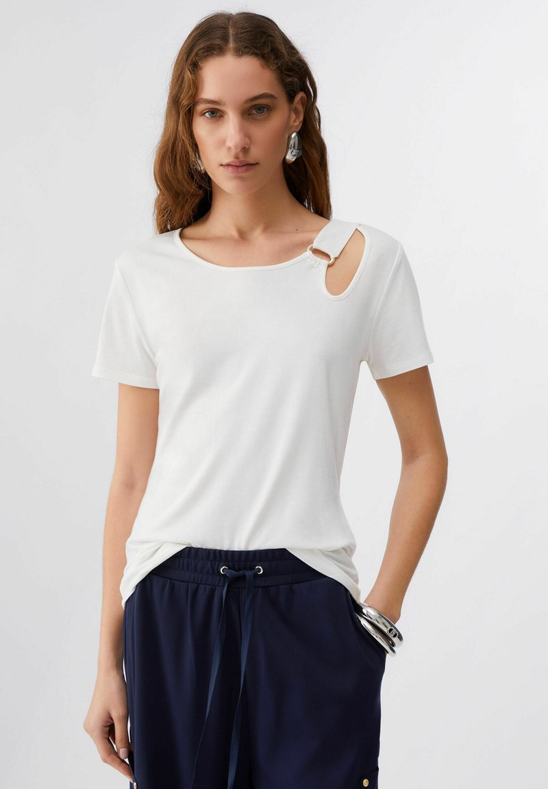 T-shirt blanc à manches courtes avec un col rond et une découpe unique à l'épaule, agrémentée d'un détail doré. Tissu lisse.