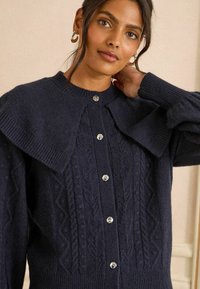Námořnický pletený cardigan s texturovaným vzorem, velkým límcem a stříbrnými knoflíky na přední straně. Měkký, útulný materiál s žebrovanými manžetami.