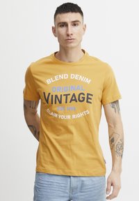 Geltona medvilninė T-shirt su apvalia iškirpte, su mėlyna ir juoda grafika „BLEND DENIM ORIGINAL VINTAGE“, dėvima su šviesiais mėlynais šortais.