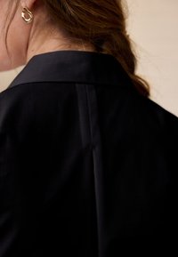 Černá košile s klasickým límečkem, s jemným leskem a čistými liniemi. Významný detail: jeden zadní skládání a zlatý kruhový náušník.