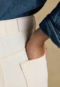 Mână alunecând în buzunarul din față al pantalonilor de culoare crem, purtați cu o jachetă de denim albastru, pe un fundal bej.