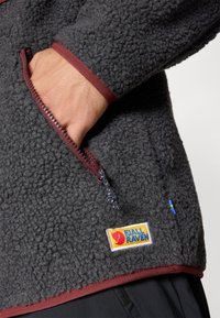 Fjällräven polárpuli sötét szürke színben, bordó szegéllyel, cipzáras zsebbel és logóval. Textúrált anyag, puha megjelenéssel.