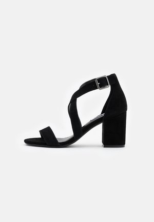 Black suede high-heeled sandal featuring a block heel, open-toed design, and cross-over strap with a buckle for secure fastening.