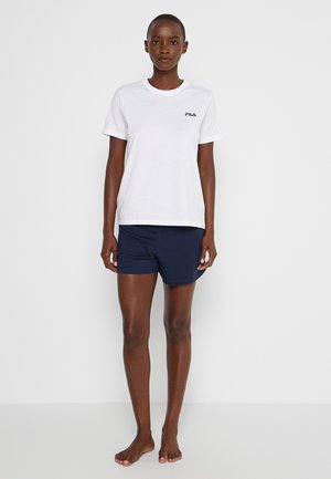Femme debout pieds nus, vêtue d'un t-shirt blanc FILA et d'un short bleu marine sur un fond uni clair.