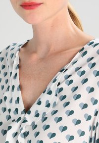 Blouse featuring a v-neck design with a white background and a pattern of small, teal and blue hearts. Lightweight, soft texture.