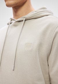Huppari vaaleasta beigeistä materiaalista, jossa on textuuripinta, nauhat ja neliön muotoinen beige logo -laatta rinnassa. Hupparissa on kengurupussimainen tasku.