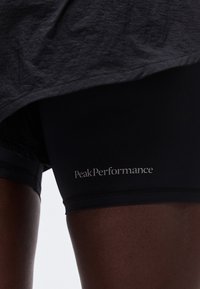 Κοντινό πλάνο ενός ατόμου που φοράει μαύρη βερμούδα με το λογότυπο "Peak Performance" στον μηρό και σκούρο γκρι πουκάμισο που καλύπτει μερικώς τη βερμούδα.