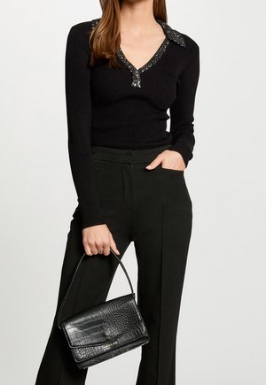 Frau trägt ein schwarzes Langarmoberteil mit Paillettenkragen und schwarze Hose, hält eine schwarze strukturierte Ledertasche mit Henkel oben.
