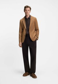 Blazer marrone sopra una polo scura, abbinato a pantaloni scuri e scarpe stringate marroni. Il blazer presenta rever a punta e tasche frontali.