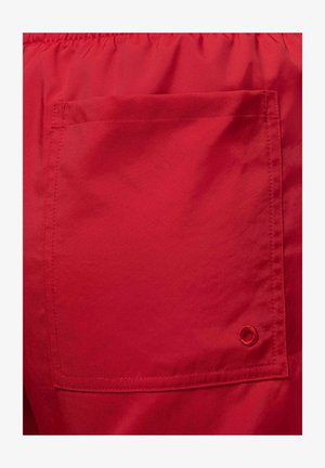 Červená tkanina s hladkým povrchom, obsahujúca štvorcovú náprsné vrecko a malý vyšívaný akcent v tvare očká. Viditeľný elastický pás.