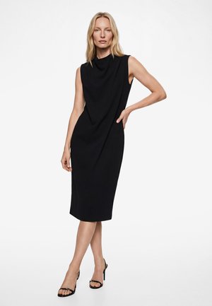 Frau in einem ärmellosen schwarzen Midikleid mit hohem Ausschnitt und schwarzen, Riemchen-Sandaletten mit hohen Absätzen vor weißem Hintergrund.