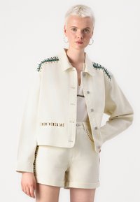 Chaqueta de mezclilla blanca con costuras decorativas en verde y dorado en los hombros y bolsillos, combinada con pantalones cortos de cintura alta a juego.